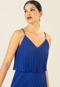 Faltiges blaues Kleid mit V-Ausschnitt und schmalen Trägern, das goldene Akzente an den Schultern sowie eine leichte, fließende Stofftextur aufweist.