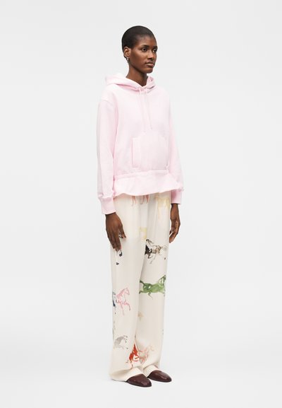 Naine, kes kannab heleroosat kapuutsiga pusa ja laia lõikega kreemikaid pükse, millel on mitmevärvilised hobusemustrid, seisab valge tausta ees.