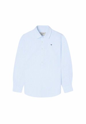 Niño con gorra de béisbol verde oliva, camisa blanca de manga larga con botones, pantalones cargo negros y zapatillas multicolor, de pie con una mano en el bolsillo.