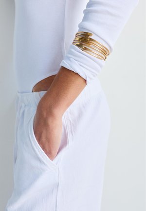 Ręka w białej bluzce z długim rękawem i spodniach z ręką w kieszeni, nosząca kilka cienkich złotych bransoletek na przedramieniu.