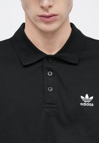 Must musta polo särk tekstureeritud kangaga, klassikalise kraega, kolmekordse nööbistikuga ja valge Adidas logo vasakul rinnal.