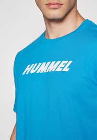 Camiseta de algodón turquesa con el logo blanco de "HUMMEL" en el pecho, mangas cortas y escote redondo. Textura suave.