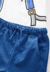 White t-shirt featuring blue suspenders graphic; navy blue cotton shorts with elastic waistband and drawstring, side pocket detail.