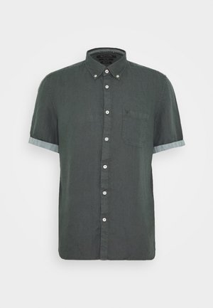 Camicia verde scuro a maniche corte con bottoni, taschino sul petto e polsini arrotolati, mostrata su uno sfondo chiaro e uniforme.