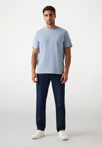 Hombre con una camiseta de manga corta azul claro, jeans azul oscuro y zapatillas blancas, de pie contra un fondo blanco liso.