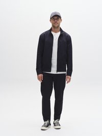 Tmavě modrá sportovní bunda a kalhoty s bílým tričkem pod nimi. Šedá čepice s vyšitým logem. Černé tenisky s bílými tkaničkami.