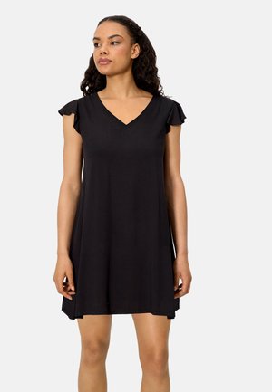 Mujer con cabello rizado que lleva un vestido negro corto con escote en V y pequeñas mangas acampanadas, de pie frente a un fondo liso y claro.