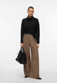 Czarny sweter z golfem, szerokie beżowe spodnie z zakładkami, czarny pasek, trzymając czarną torbę tote. Gładkie tekstury i zharmonizowane kształty.