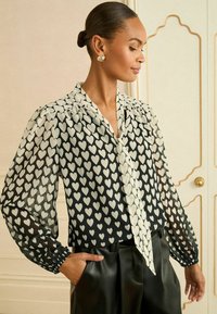 Blusa nera trasparente con motivo a cuori bianchi, caratterizzata da un colletto con lacci e maniche a sbuffo. Abbinata a pantaloni neri a vita alta.