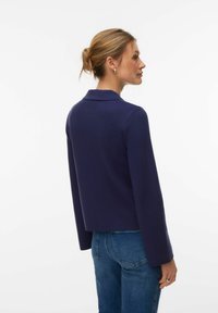 Veste courte bleu marine avec col et manches longues, confectionnée en tissu texturé. Portée avec un jean en denim, dévoilant un design ajusté au dos.