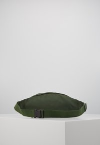 Marsupio verde oliva realizzato in tessuto liscio, caratterizzato da una forma curvata, una tracolla regolabile e una fibbia in plastica nera.
