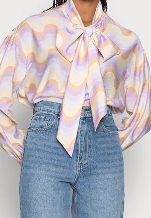 Person trägt hoch taillierte blaue Jeans und eine langärmlige Bluse mit pastellfarbenen wellenförmigen Mustern und einer großen gebundenen Schleife am Hals.