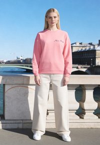 Jeune femme portant un sweat-shirt rose "Mira Paris" et un pantalon blanc large, debout auprès d'une rambarde en pierre au bord de la rivière sous un ciel bleu clair.