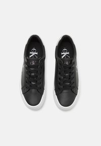 Paire de baskets noires basses avec semelles blanches, lacets noirs et logos "CK" blancs sur les côtés et les semelles intérieures, vues de dessus.