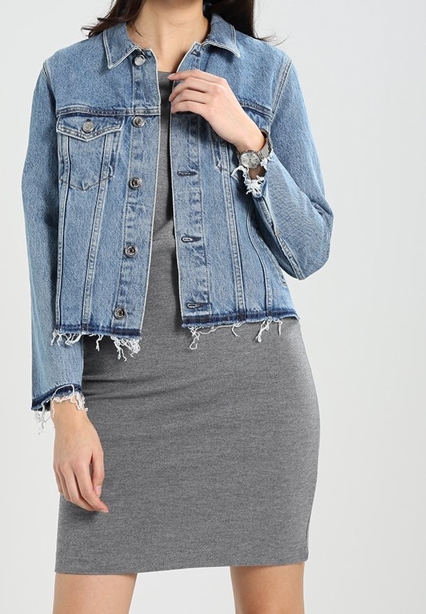 Kurtka dżinsowa, jasnoniebieska, krótkiego kroju z postrzępionym dołem i przednimi kieszeniami, nałożona na dopasowaną szarą sukienkę, ręce częściowo widoczne.