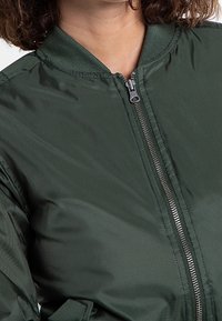 Zielona kurtka bombowiec wykonana z gładkiego nylonu, z zamkiem błyskawicznym z przodu, prążkowanym kołnierzem i bez widocznych wzorów czy akcentów.