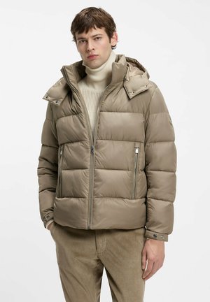 Young man wearing tan puffer jacket, cream turtleneck sweater, and brown corduroy pants, standing with one hand in pocket.