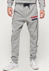 Grijze sweatpants met elastische tailleband en trekkoord. Heeft rode en marineblauwe borduursels "SUPERDRY" en "DRY" op de linker bovenbeen. Zijkanten met strepen.