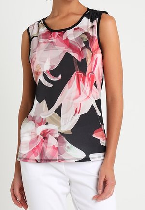 Haut noir sans manches avec un grand imprimé floral rose et blanc porté avec un pantalon blanc sur une personne debout devant un fond uni.
