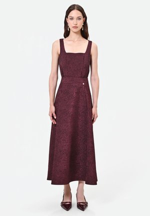 Femme portant une robe midi sans manches texturée violet foncé avec un col carré et des chaussures pointues assorties, debout devant un fond uni.