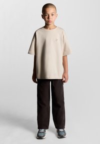 Beige oversized t-shirt med et lille broderet design, parret med løse sorte bukser og grå sneakers med blå accenter.