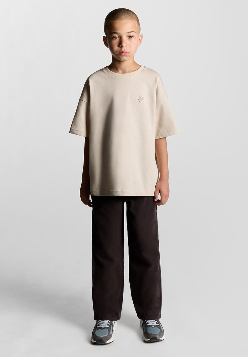 Beige oversized t-shirt med et lille broderet design, parret med løse sorte bukser og grå sneakers med blå accenter.