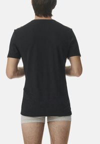 Schwarzes T-Shirt mit kurzen Ärmeln aus Baumwolle, mit einem Rundhalsausschnitt und einer taillierten Passform, von hinten betrachtet. Darunter helle enganliegende Shorts.