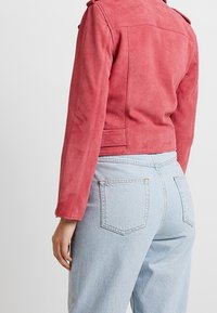 Dopasowana czerwona zamszowa kurtka z długimi rękawami, pagonami na ramionach i krótkim krojem, w połączeniu z jasnoniebieskimi, jeansami z wysokim stanem.