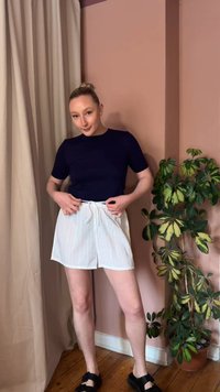 Γυναίκα που ρυθμίζει λευκά ριγέ σορτς, φοράει μαύρο τοπ με κοντά μανίκια, στέκεται σε ξύλινο πάτωμα κοντά σε ροζ τοίχο και πράσινο κασπώ με φυτό.