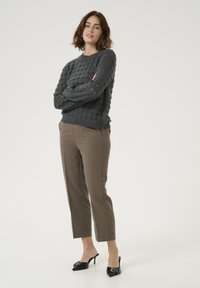 Maglione grigio con lavorazione a maglia grossa e tessitura aperta, abbinato a pantaloni beige a sigaretta e mules neri con tacco e dettagli decorativi.