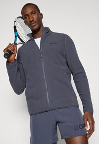 Vīriešu pelēks flīsa jaka ar rāvējslēdzēja aizdari, ribotiem piedodamiem un apkakli. Apvienota ar pelēkiem šortiem, kuros ir logotipa prints. Tur rokās tenisa raketi.