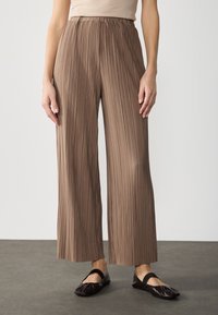 Bruine plissé wijde pijpen broek met een elastische tailleband, voorzien van een verticale geribbelde structuur en een vloeiende stof. Gecombineerd met zwarte ballerina's.