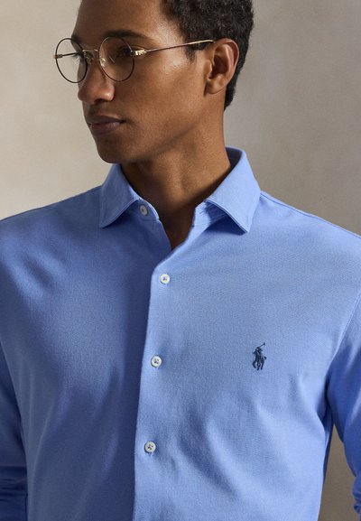 Άνδρας που φορά στρογγυλά γυαλιά και ανοιχτό μπλε πουκάμισο με κουμπιά, με μικρό σκούρο κεντητό λογότυπο αθλητή πόλο στο στήθος.