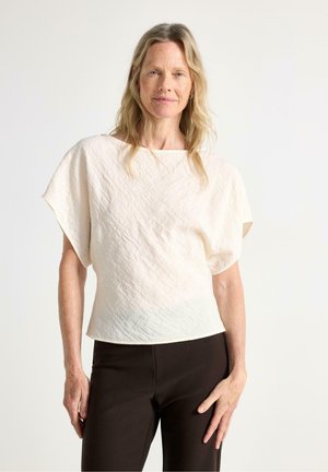 Donna con capelli biondo chiaro che indossa una maglia testurizzata bianco sporco a maniche corte e pantaloni neri, in piedi davanti a uno sfondo chiaro semplice.