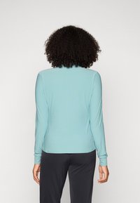 Long-sleeve top in light blue fabric with a smooth texture. Features a rounded hem and fitted cuffs, paired with dark pants.