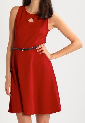 Femme portant une robe rouge sans manches avec une petite ouverture devant et une fine ceinture noire, debout, la main sur la hanche, sur un fond uni.