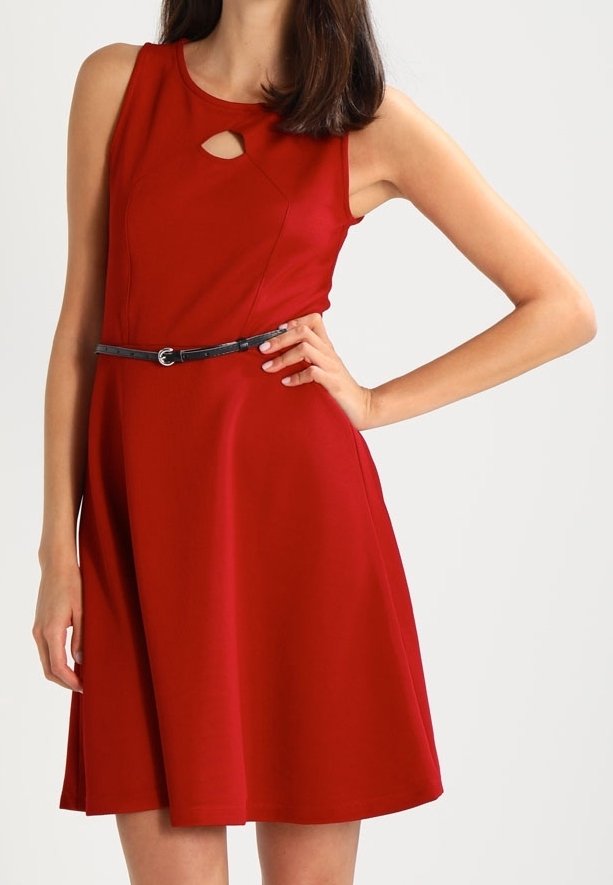 Femme portant une robe rouge sans manches avec une petite ouverture devant et une fine ceinture noire, debout, la main sur la hanche, sur un fond uni.