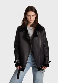 Black leather jacket with a textured finish, featuring a shearling collar, zip closure, and adjustable straps at the cuff.