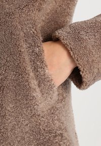 Ręka włożona w kieszeń brązowego, fakturowanego, puszystego rękawa kurtki z polaru na neutralnym tle.