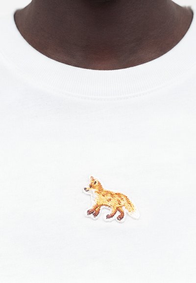 Άτομο με σκουρόχρωμο δέρμα που φοράει λευκό μπλουζάκι με στρογγυλή λαιμόκοψη και μικρό κεντητό πορτοκαλί αλεπού στο κέντρο του στήθους.