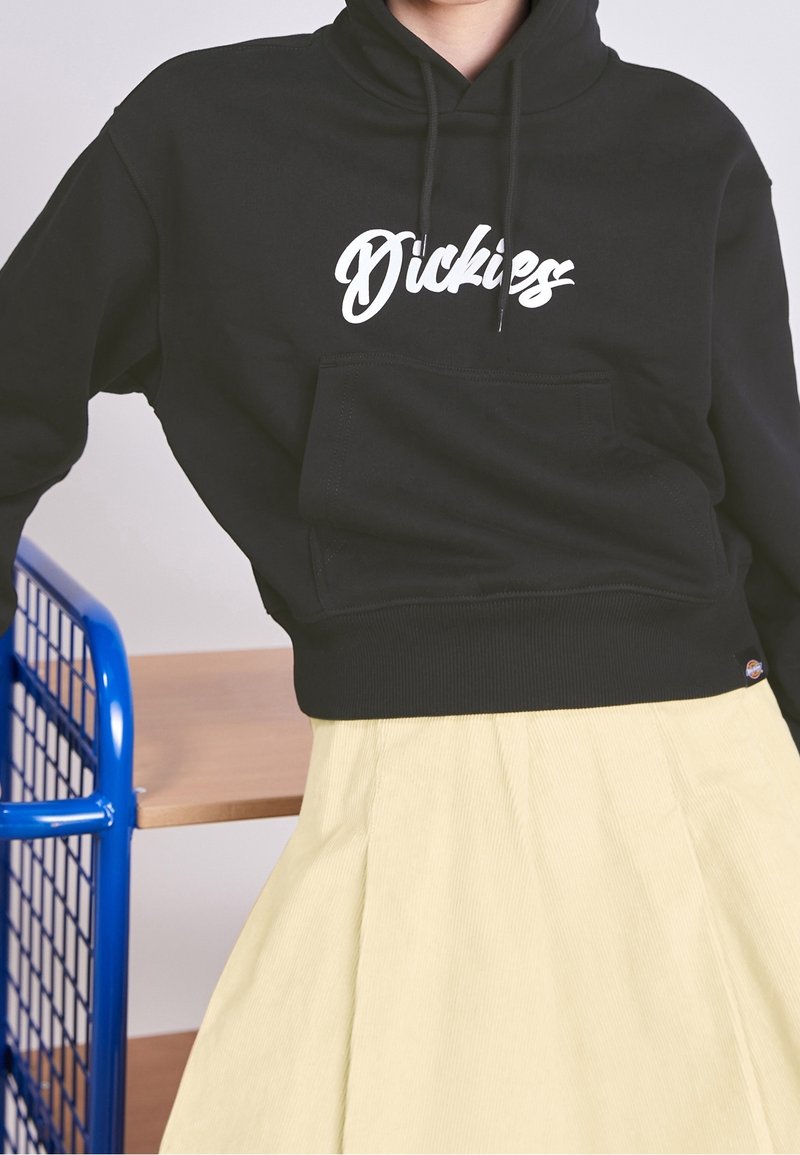 Personne portant un sweat à capuche Dickies noir avec une poche avant et une jupe jaune clair, appuyée contre une structure en métal et bois bleue.