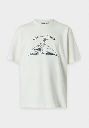 Giovane uomo che indossa una T-shirt bianca con il testo e la grafica "Ride Your Iceberg", jeans neri larghi, sneakers bianche, in piedi con una mano in tasca.