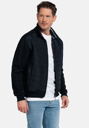 Veste bomber noire avec un devant matelassé, des panneaux de manches en tricot et des poches zippées. Elle possède un col montant et est portée sur une chemise blanche.