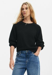 Maglione nero lavorato a maglia con dettagli a trecce sulle maniche, scollatura rotonda e vestibilità rilassata, abbinato a jeans blu a vita alta.