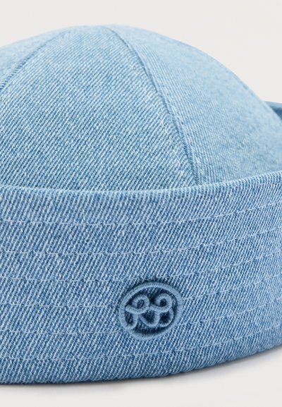 Μπλε denim καπέλο τύπου κουβά με ραμμένο κυκλικό έμβλημα στο πλάι, με διακριτικό υφασμάτινο υφή και ορατές γραμμές ραφής.