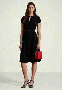 Egy nő fekete övvel ellátott, térd fölé érő ruhát visel, fekete magassarkú szandált hord, és egy kis piros kézitáskát tart, egy sima háttér előtt áll.