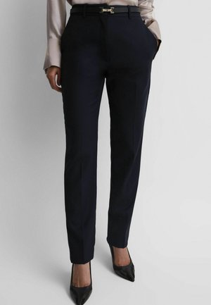 Vrouw draagt op maat gemaakte zwarte broek met een zwarte riem, beige blouse en zwarte puntige hoge hakken, staand met handen in de zakken.