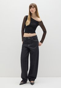 Top nero a spalla unica con maniche lunghe trasparenti, abbinato a pantaloni larghi in denim scuro con cuciture visibili e un fit oversize.