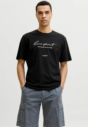 Mand med kort krøllet hår iført sort t-shirt med teksten "Core Sport Worldwide Since 1990" og grå cargoshorts, hænderne i lommerne, neutral baggrund.