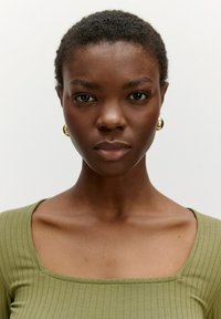 Model trägt ein ribbed, olivgrünes Oberteil mit einem eckigen Ausschnitt. Goldene Creolen betonen den Look vor einem schlichten Hintergrund.